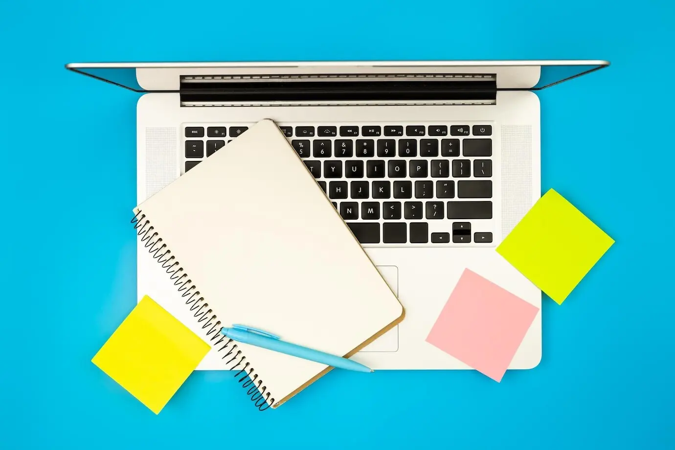 Laptop, Notizblock und Erinnerungszettel aus Papier auf blauem Hintergrund, Draufsicht.
